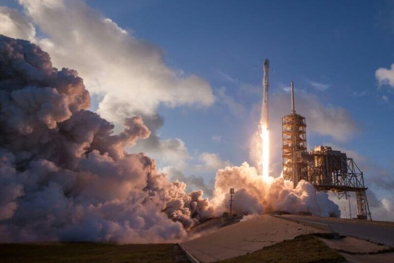 Image of a powerful rocket in the midst of a fiery and dynamic launch at Kennedy Space Center, with billowing clouds of smoke and intense flames at the base. The rocket is set against a clear blue sky, symbolizing the beginning of a momentous journey, and reflecting the ambitious and forward-moving spirit of the website's mission in making IT concepts accessible and engaging.